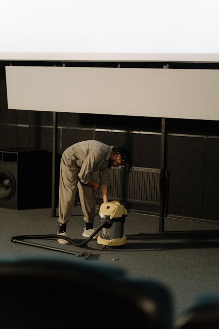 A person is vacuuming a patterned carpet in a well-lit room, with only their white sneakers and part of their legs visible. The vacuum cleaner has a black body and a long, black suction tube connected