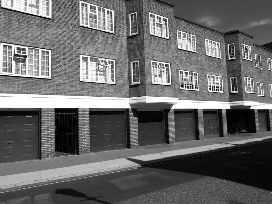 Photograph of a modern multi-storey residential building with a beige brick facade and numerous uniform windows, situated on Stockwell Park Estate SW9. The image features surrounding green trees with