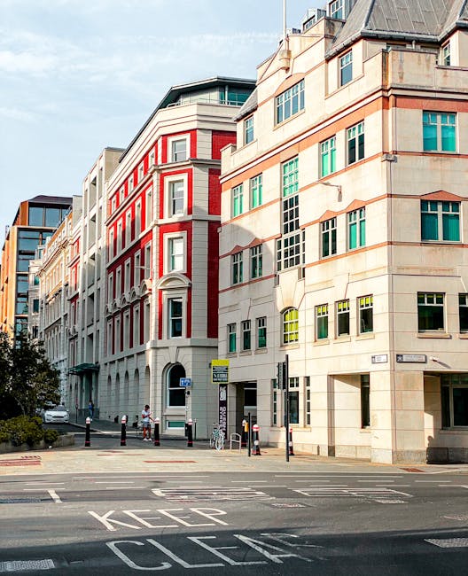 Image of a city street with modern multi-story office buildings featuring large glass windows and mixed materials including beige stone and red brick accents. The street has a 'KEEP CLEAR' marking on 